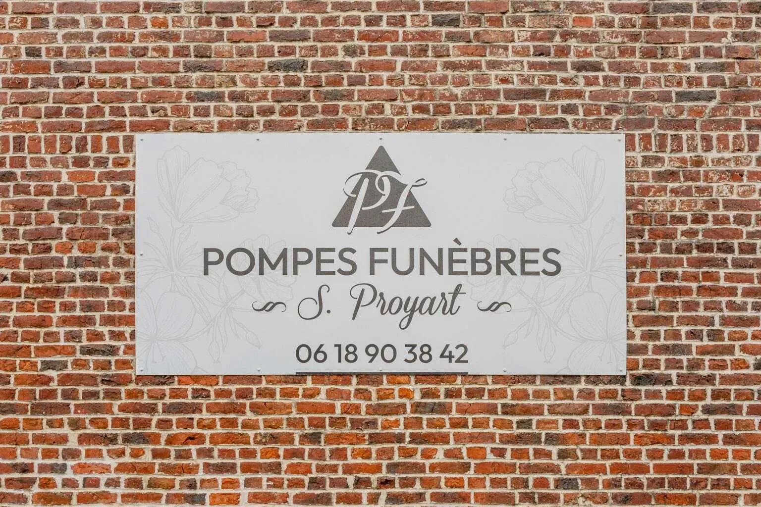 Plaque des Pompes Funèbres S. Proyart fixée sur un mur en briques rouges.
